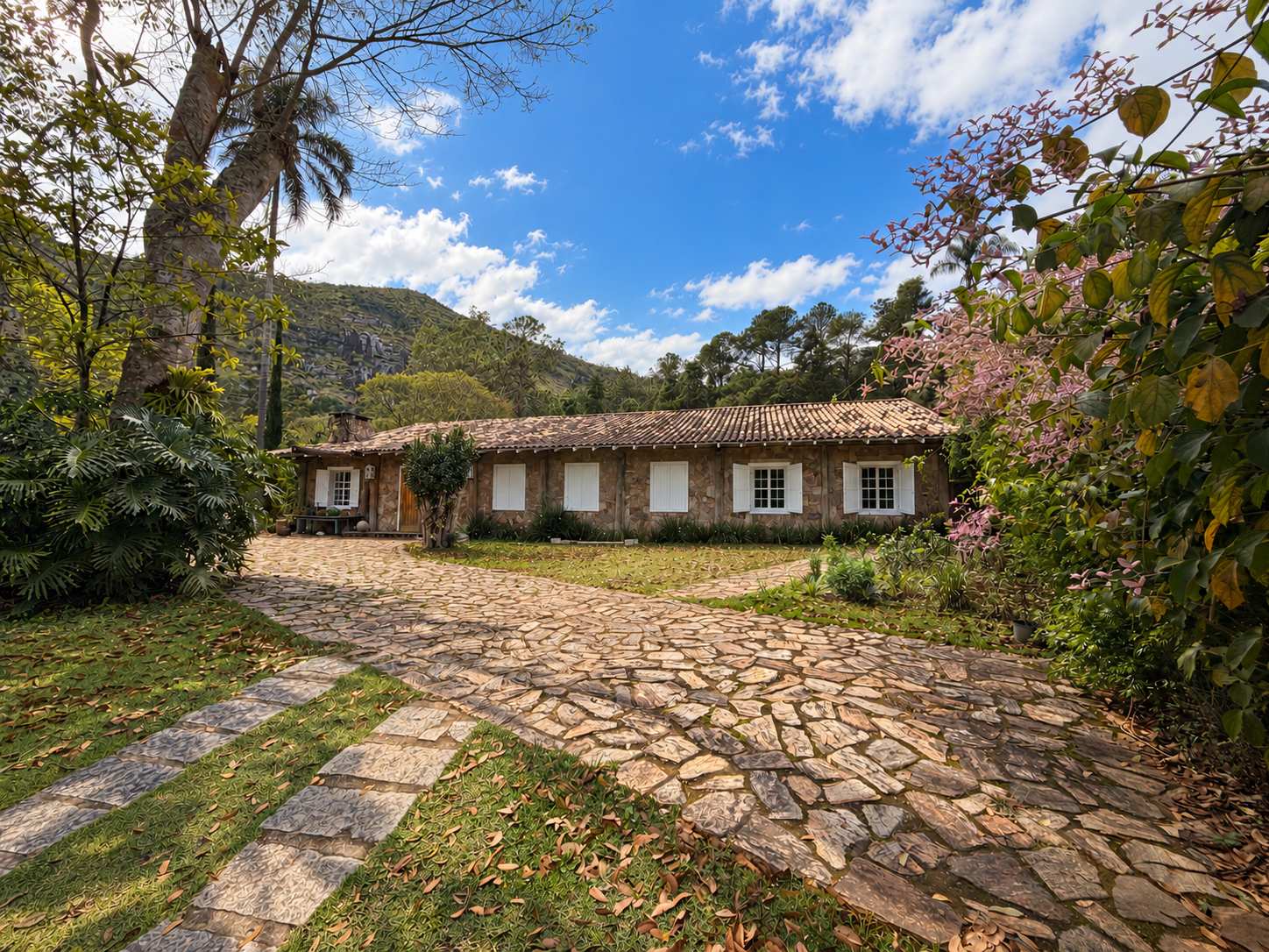 Fachada da Pousada FAVO: casa térrea de pedra com telhado de barro e janelas de venezianas brancas, pátio de pedra à frente e a serra de Petrópolis ao fundo sob céu azul