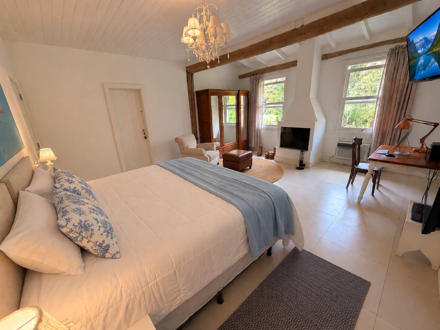 Suíte 01 de dia, em plano aberto: cama king com manta azul-clara, lustre, vigas de madeira, lareira, poltrona, armário e escrivaninha junto às janelas para o verde