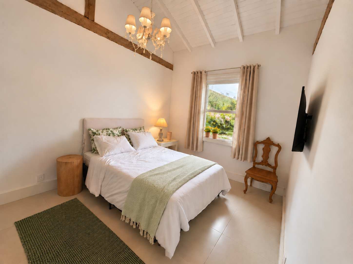 Suíte 02 em plano aberto: cama de casal com manta verde-clara, lustre de cúpulas, viga de madeira no pé-direito alto, janela com cortinas e vista para o morro, cadeira de madeira e TV na parede