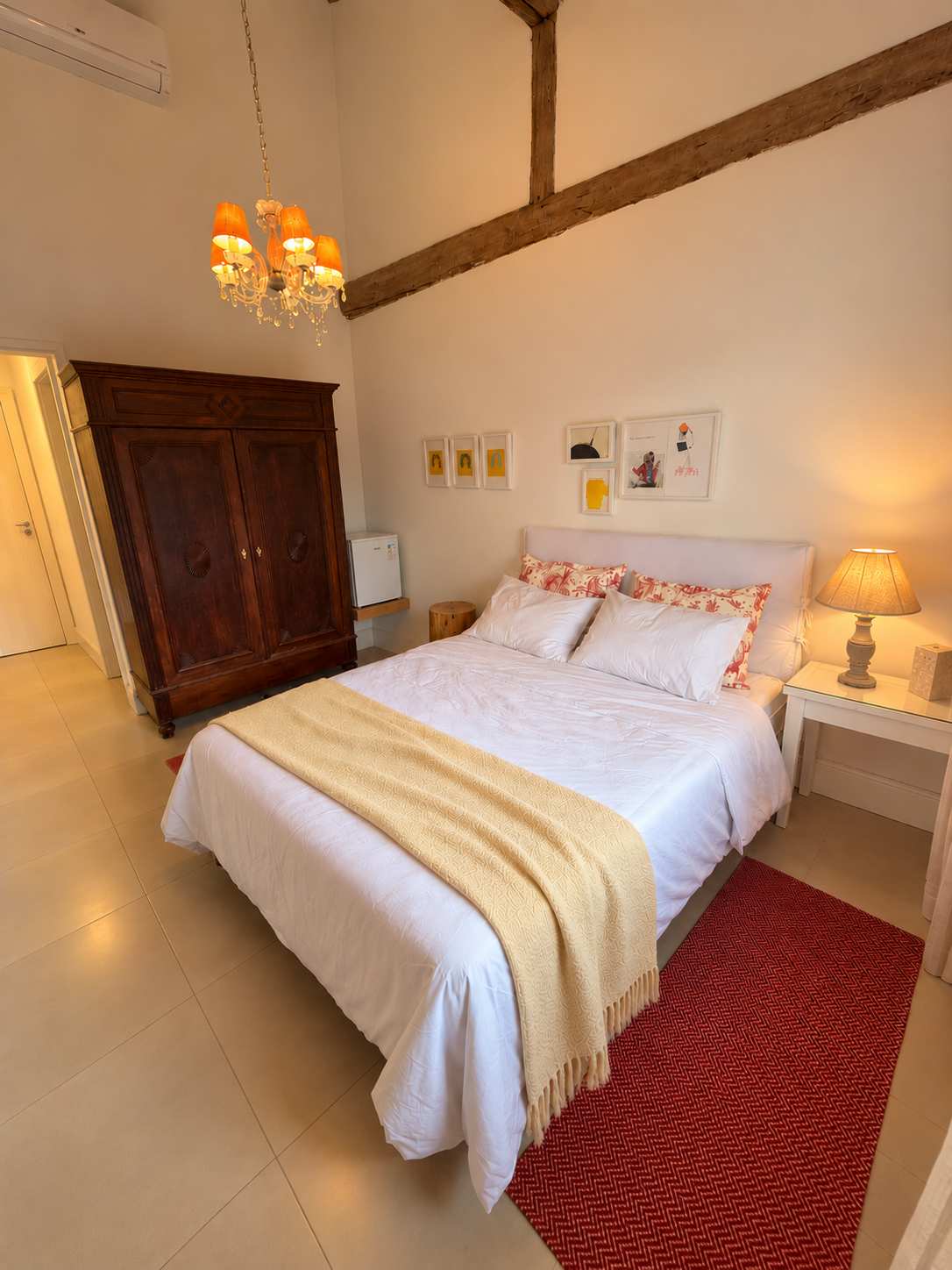 Suíte 04 vista da cama: armário de madeira maciça, frigobar e quadros na parede branca sob as vigas